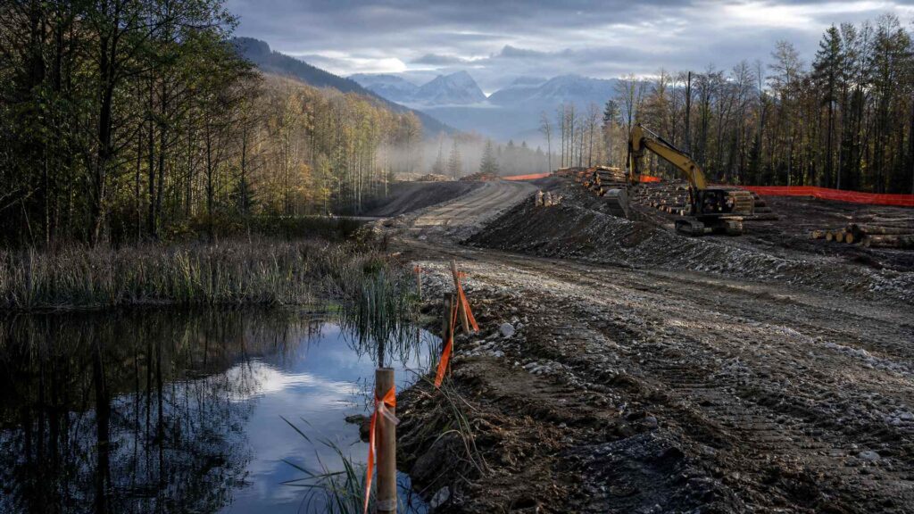 Image éditoriale générée par IA, non documentaire : chantier de l'A412 dans le Bas-Chablais ; à gauche, une zone humide intacte avec roseaux, eau sombre et rubans de balisage ; au centre, une plateforme de remblai en gravier et des ornières ; à droite, des troncs abattus, une pelle mécanique jaune et des engins de terrassement ; collines boisées et Alpes dans la brume au fond.
