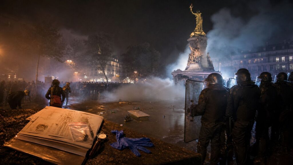 Photo de nuit place de la République : au premier plan, sur un muret de pierre mouillée, un dossier de médecine légale ouvert avec ses schémas, une montre arrêtée et un gant en nitrile ; à droite, une ligne de policiers casqués, visières baissées, boucliers levés ; à gauche, une foule qui recule dans un nuage de gaz ; la statue de la République domine la place dans la fumée éclairée.