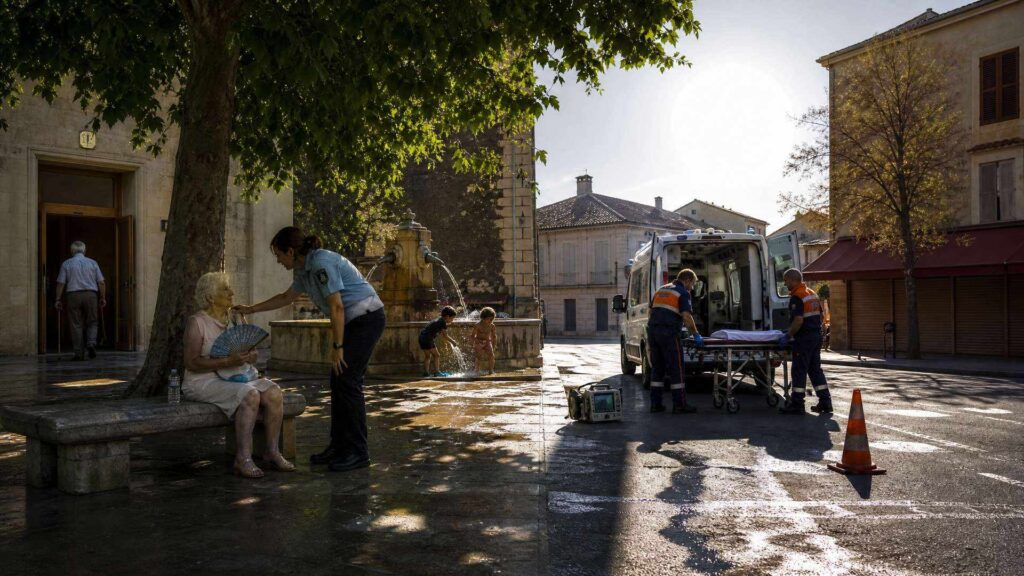 Sur une place de ville du Sud écrasée de soleil, une femme âgée s'éventaille sur un banc de pierre à l'ombre d'un platane tandis qu'une agente se penche vers elle ; à droite, en plein soleil, deux secouristes déploient un brancard près d'une ambulance, devant une fontaine où jouent des enfants.