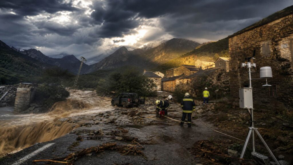 Route départementale corse coupée par un torrent d'eau brune au petit matin : culée de pont effondrée et branchages sur la chaussée au premier plan, deux sapeurs-pompiers en tenue sombre et bandes réfléchissantes près d'une motopompe, un 4x4 embourbé portière ouverte, une station météorologique avec pluviomètre et anémomètre à droite, hameau de pierre aux volets clos derrière, montagne et ciel d'orage en arrière-plan.