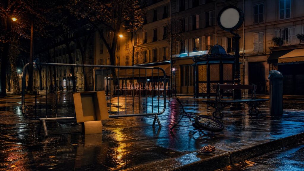 Rue parisienne deserte sous la pluie a la nuit tombee : barriere de police en travers du trottoir, chemise cartonnee ouverte et vide posee contre elle, velo d'enfant couche au sol ; au fond un banc vide, des facades aux volets tires et une horloge publique sans aiguilles.