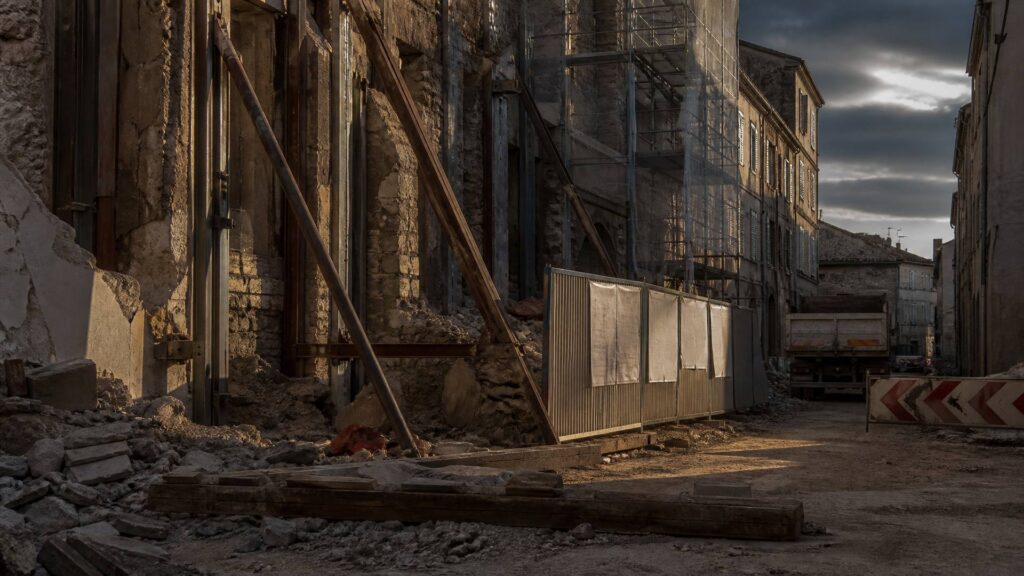 Rue en reconstruction : façade effondrée étayée, gravats au sol, échafaudage, plan de chantier punaisé sur une palissade, camion-benne au fond.