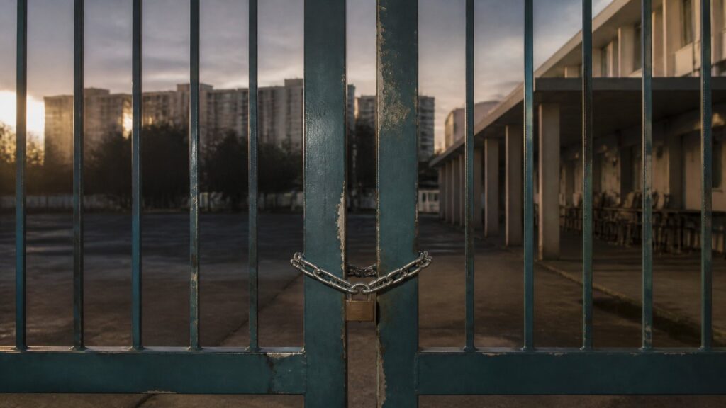 Portail d'un lycée de banlieue fermé par une chaîne et un cadenas, cour vide à l'aube