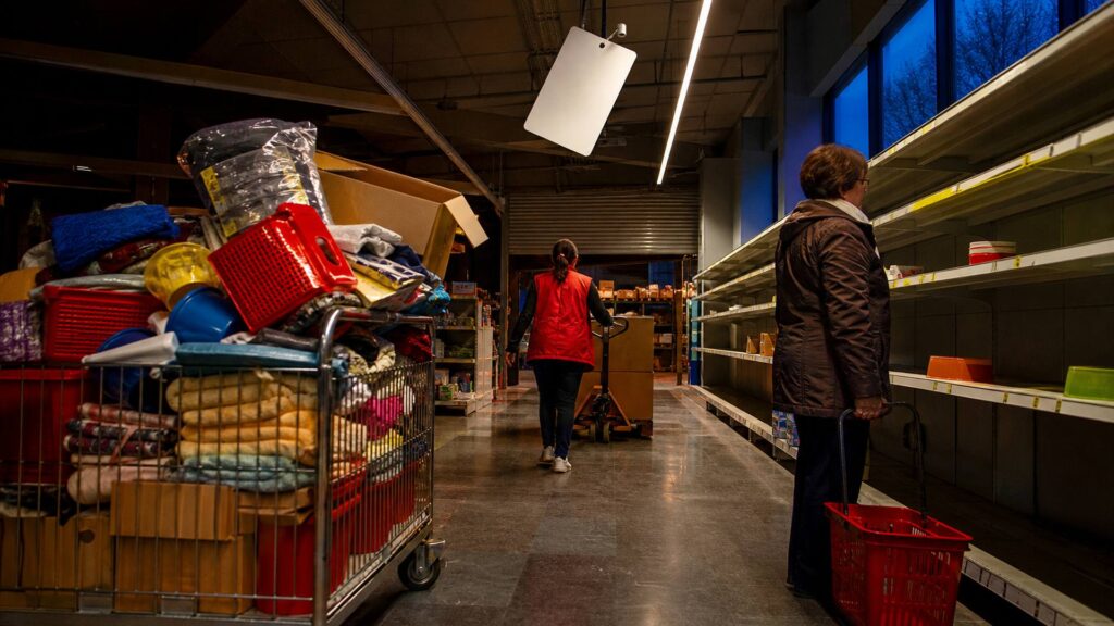 Intérieur d'un magasin de bazar discount en fin de journée : chariot chargé d'articles colorés, cartons ouverts au sol, employée en gilet rouge vue de dos poussant un transpalette, cliente âgée avec un panier, étagères à moitié vides et rideau métallique au fond.