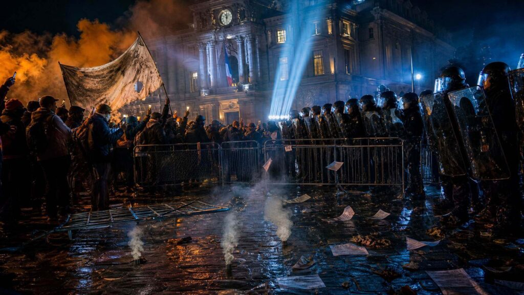 Rue française de nuit devant une préfecture monumentale : à gauche une foule de manifestants éclairée par des téléphones, à droite un rang de policiers casqués sous des projecteurs bleus, barrières renversées et douilles fumantes au sol.