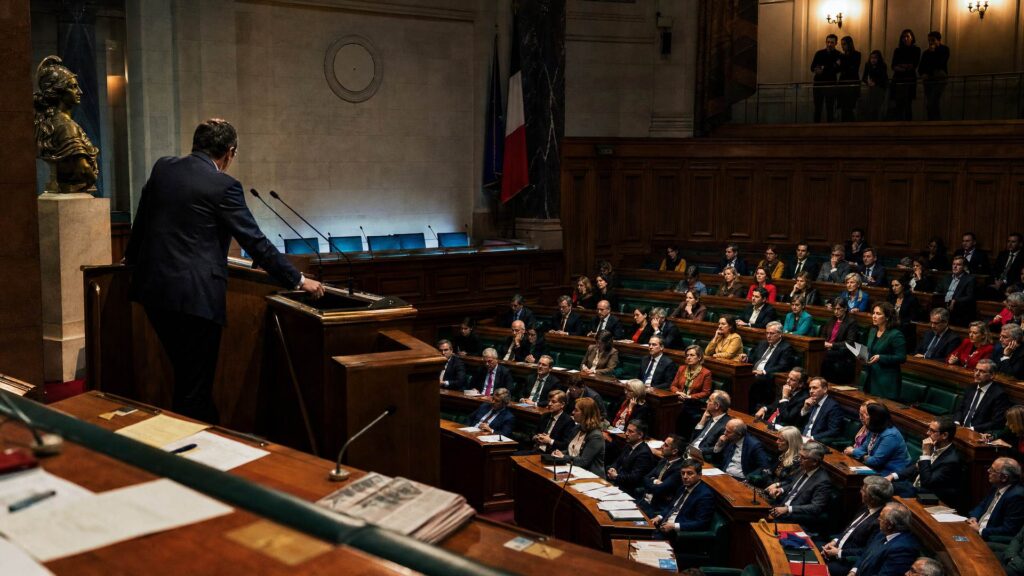 Séance de nuit à l'Assemblée nationale : un orateur vu de dos à la tribune, des députés sur les gradins verts, le public dans la galerie sous une lumière chaude, un buste de Marianne à gauche.