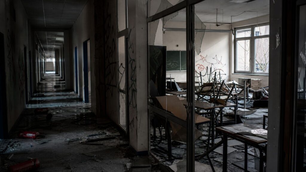 Couloir et salle de classe d’un lycée dévastés : vitres brisées, tables renversées, murs couverts de graffitis illisibles, extincteur au sol.