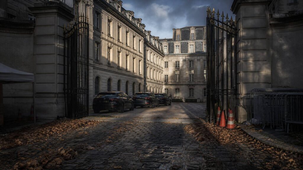 Cour d'une préfecture sous escorte : grilles monumentales, véhicules banalisés garés en file, barrières de sécurité rangées contre un mur, parvis vide.
