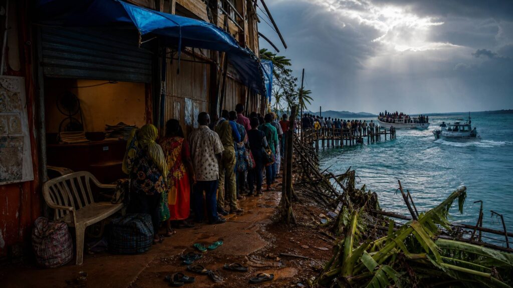 Devant un bâtiment administratif aux volets baissés, une longue file de personnes vues de dos attend sous une bâche bleue ; la queue se prolonge sur un ponton de bois vers un lagon turquoise où un bateau blanc chargé de passagers croise un canot à moteur, sous un ciel de mousson traversé d’un rayon de lumière.
