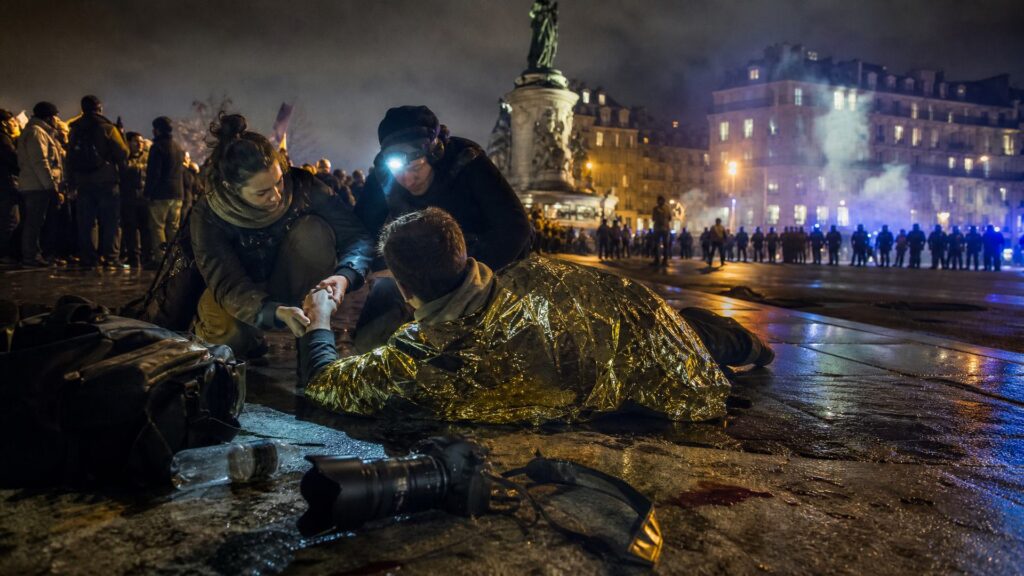 Sur les pavés mouillés d'une place parisienne, un appareil photo tombé près d'un jeune homme allongé sous une couverture de survie, entouré de deux personnes agenouillées, avec la statue de la République et un cordon de policiers en arrière-plan.
