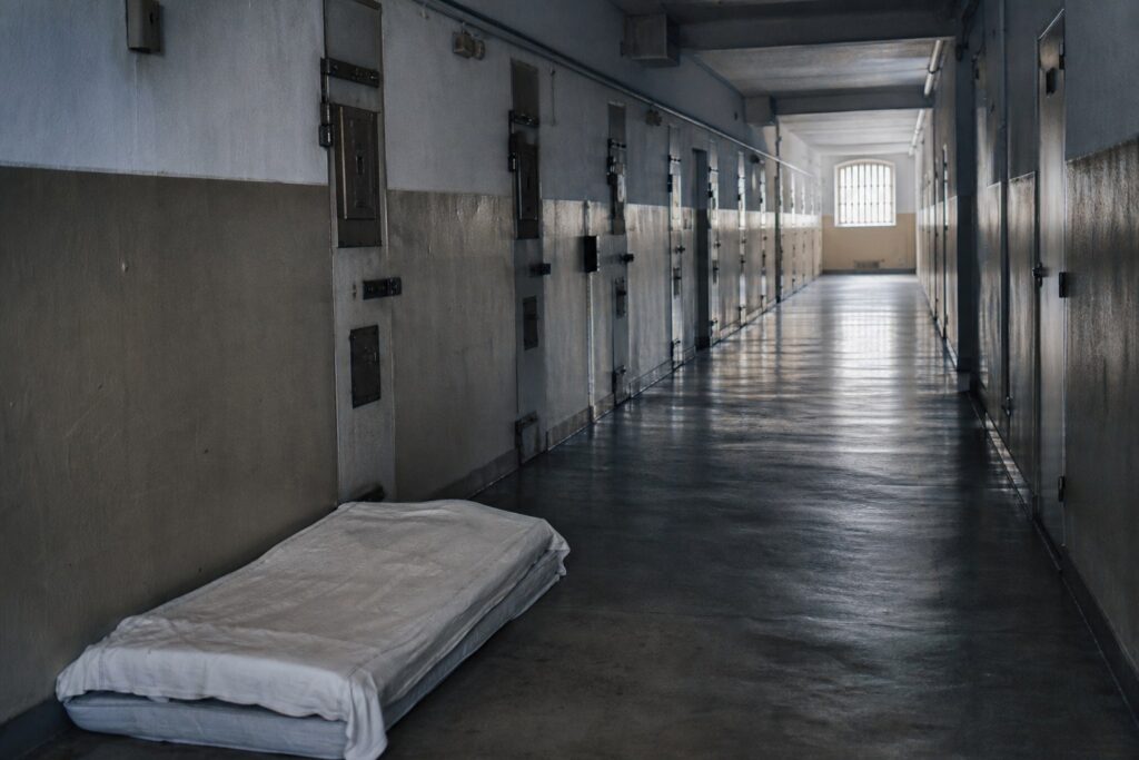 Couloir de prison faiblement éclairé, portes de cellules fermées et matelas posé au sol.