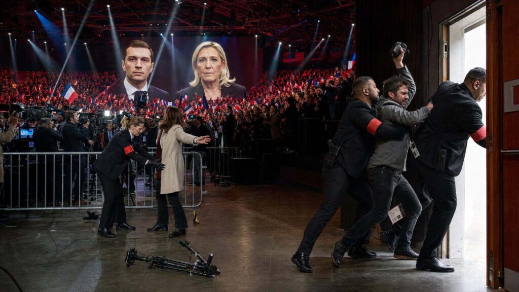 Intérieur d'une grande salle de congrès : au premier plan, deux agents de sécurité en costume sombre et brassard rouge poussent un photographe vers une porte lumineuse, un trépied renversé au sol ; derrière une barrière métallique, des journalistes et des caméras sous une lumière blanche froide ; au fond, un écran géant porte les portraits de Jordan Bardella et de Marine Le Pen, devant des milliers de militants debout agitant des drapeaux tricolores.