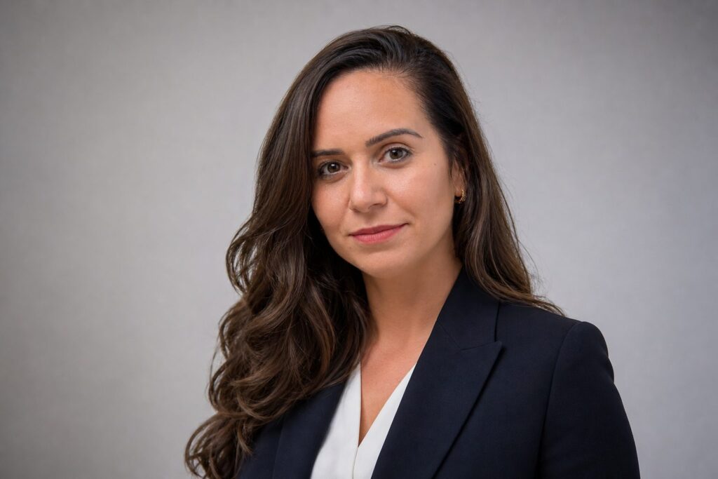 Portrait en buste d'une femme politique, cheveux longs châtain foncé, veste sombre, fond gris neutre flou.