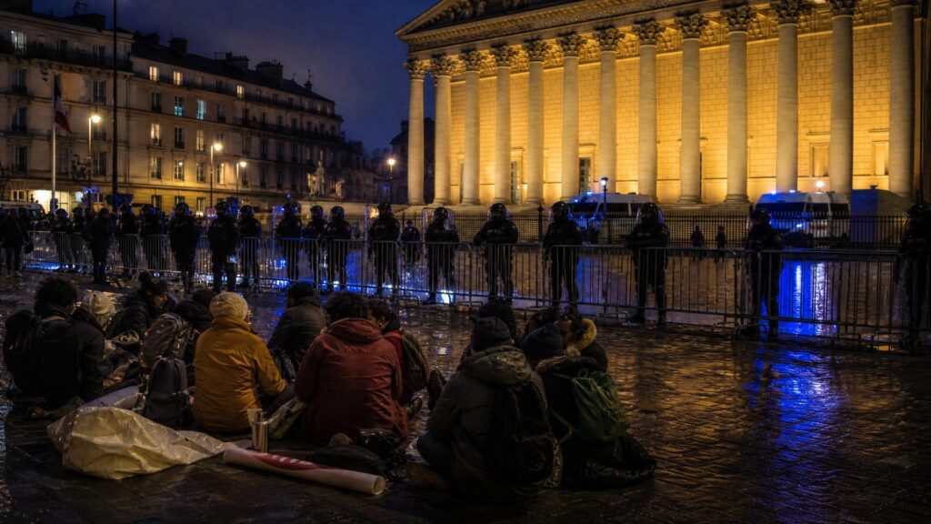 Devant la colonnade éclairée d'un palais institutionnel, des manifestants assis de dos sur le pavé, sacs et banderoles roulées à leurs côtés, face à une haie de policiers en uniforme sombre derrière des barrières métalliques, gyrophares bleus dans la nuit.