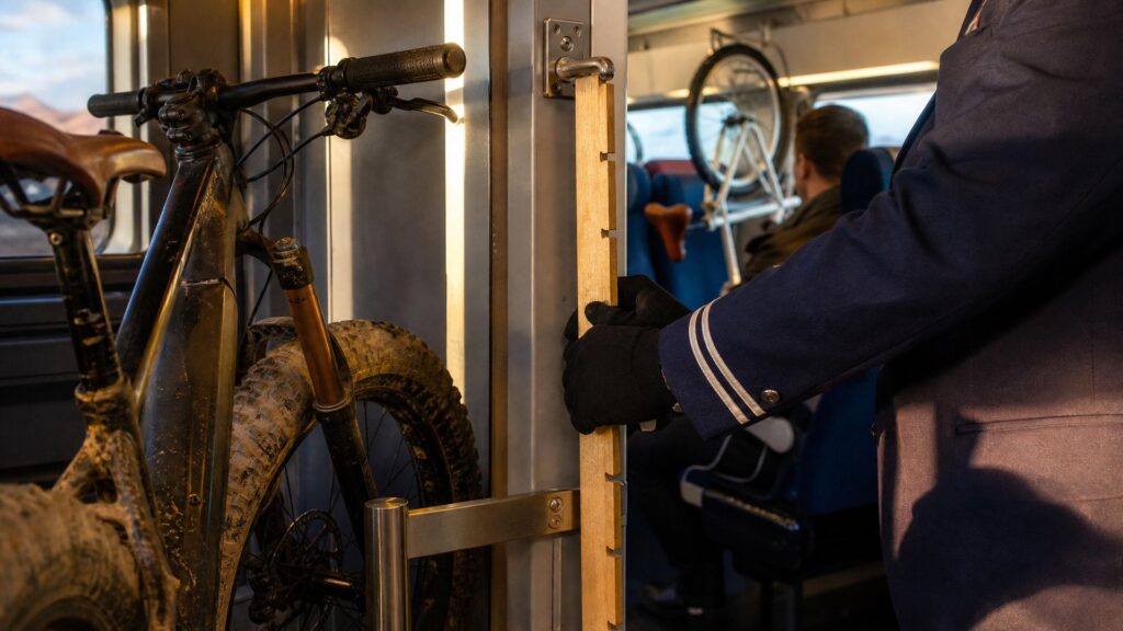 Dans l'espace vélo d'une voiture de train, un pneu de VTT large et couvert de boue est comparé à une jauge en bois clair à encoches tenue par une main gantée, sous un crochet métallique fixé au montant ; à l'arrière-plan, un voyageur assis de trois quarts dos et une roue de vélo suspendue devant des sièges bleus.