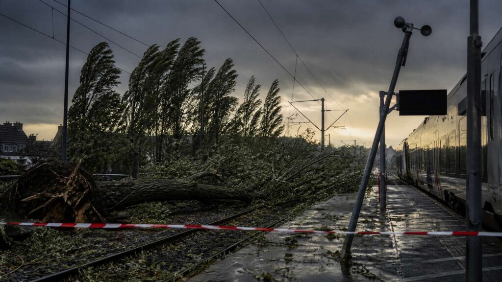 Illustration éditoriale générée par IA : quai de gare normande balayé par le vent, arbre tombé en travers des voies, caténaires secouées, anémomètre sur un mât, rame arrêtée et quai désert sous un ciel de tempête.